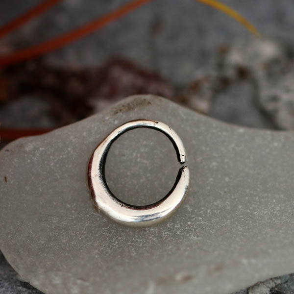 Placed on a piece of semi-translucent glass, the AMA Seamless Nose Ring in Silver by Kaya Movement features a smooth, slightly curved design. The 18 gauge 925 silver body jewelry stands out against the blurred background, with hints of earthy tones and fine, wispy plant material complementing its elegance.